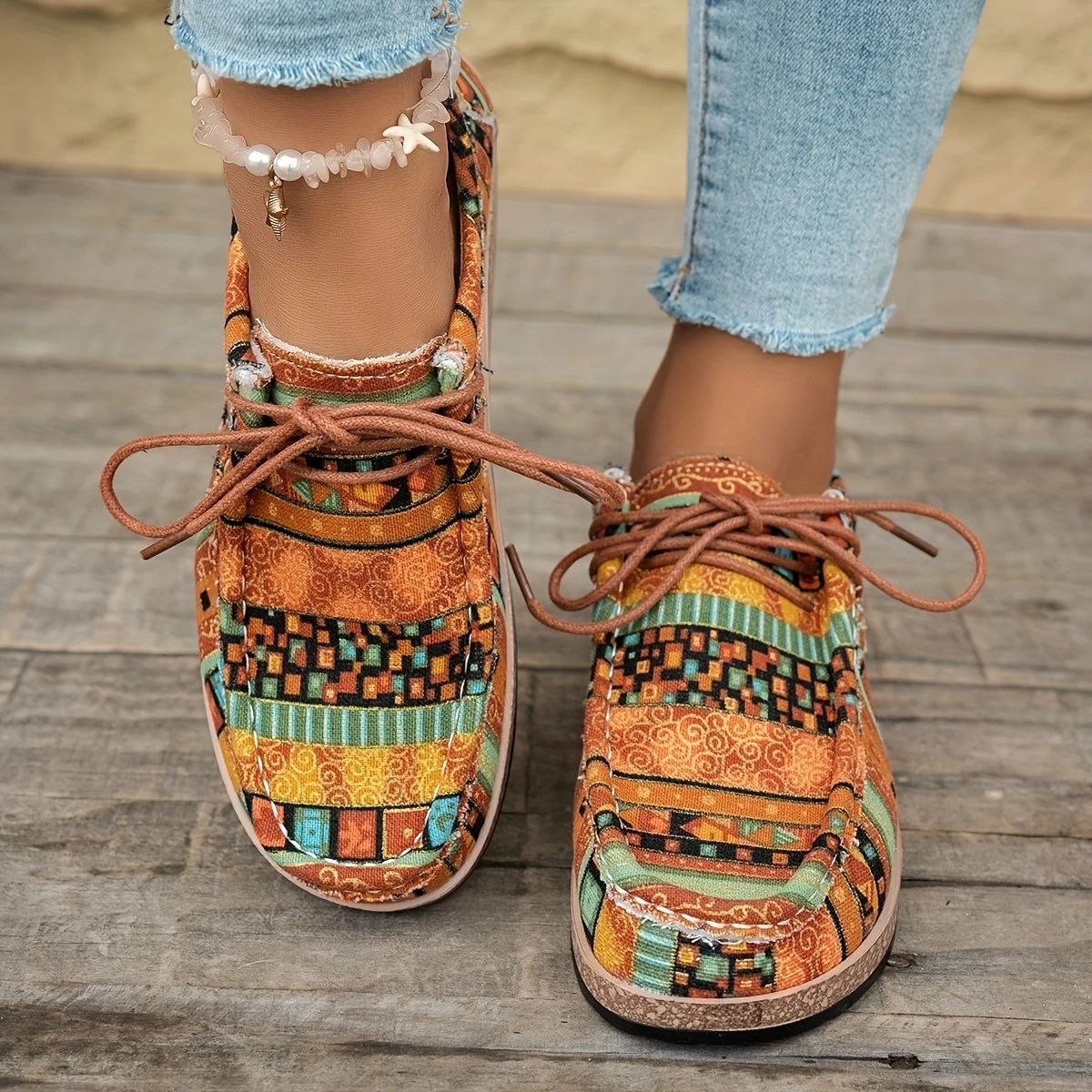 Colorful patterned casual shoes with brown laces on feet wearing light blue frayed jeans and a shell bead anklet
