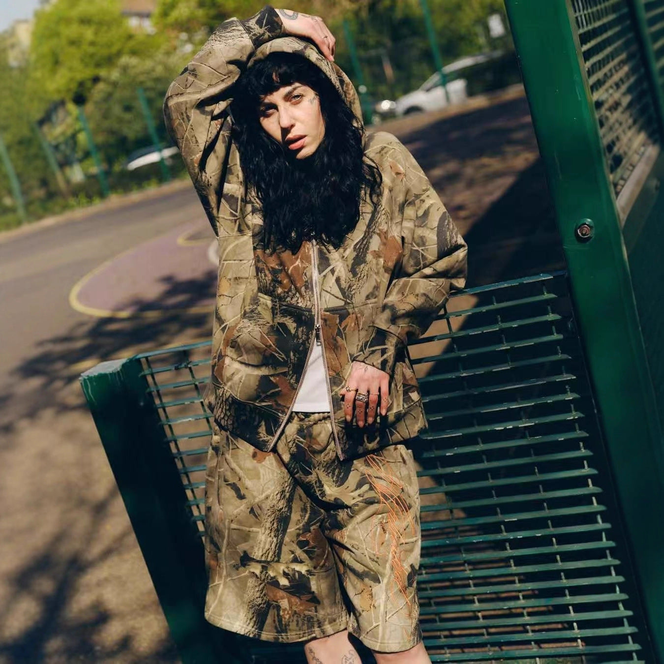 Woman in camo hoodie and matching shorts posing outdoors on green metal bench