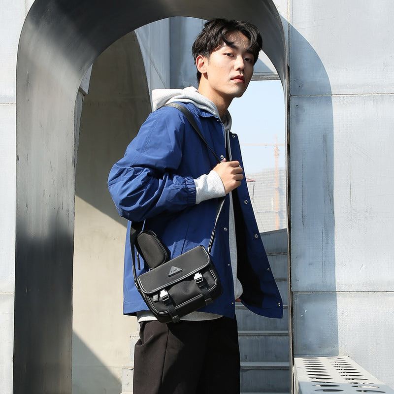 Young man wearing blue jacket and gray hoodie carrying black nylon messenger bag with strap outdoors
