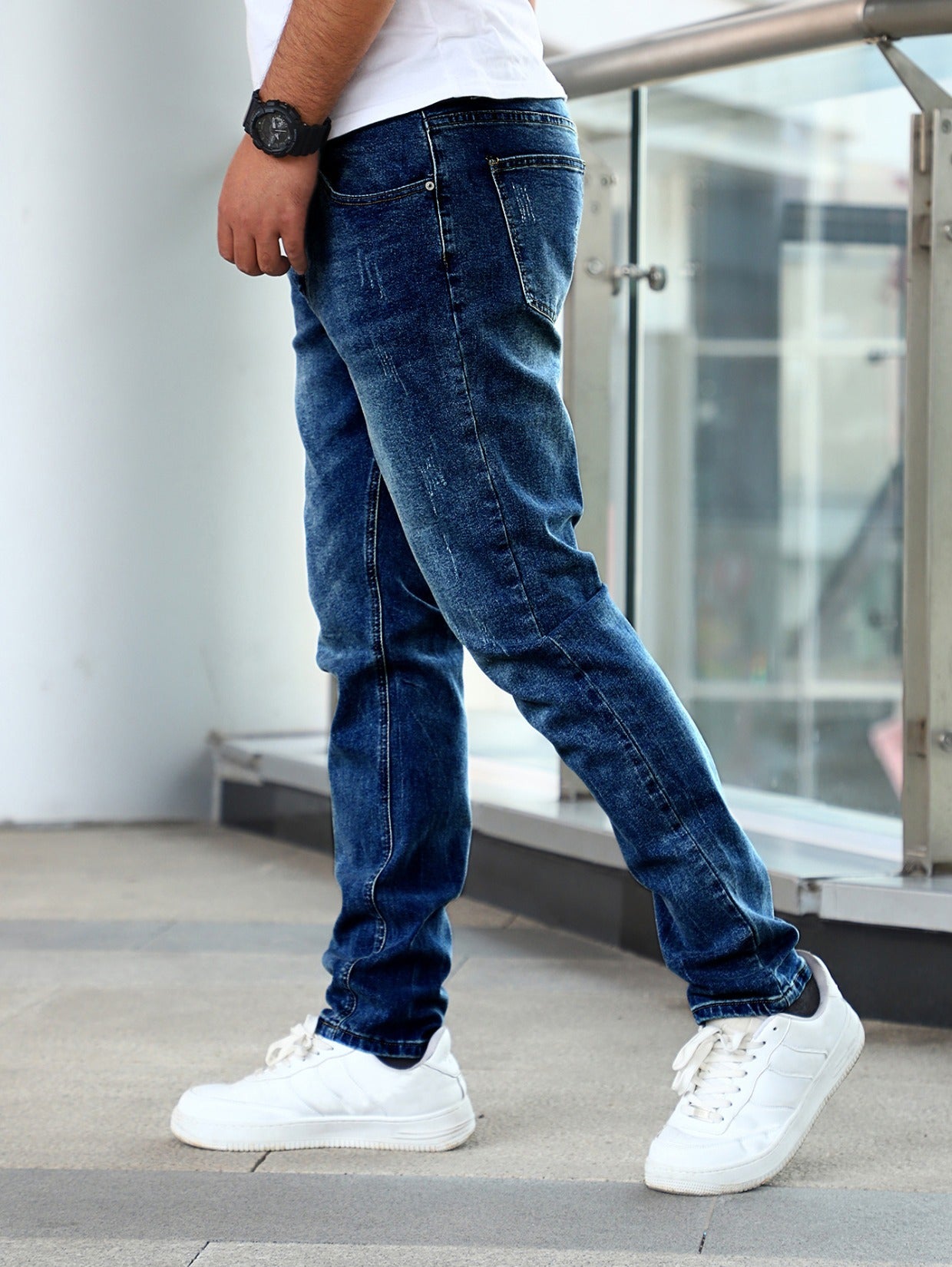 Man wearing slim fit stretch blue denim jeans and white sneakers, standing indoors near glass railing