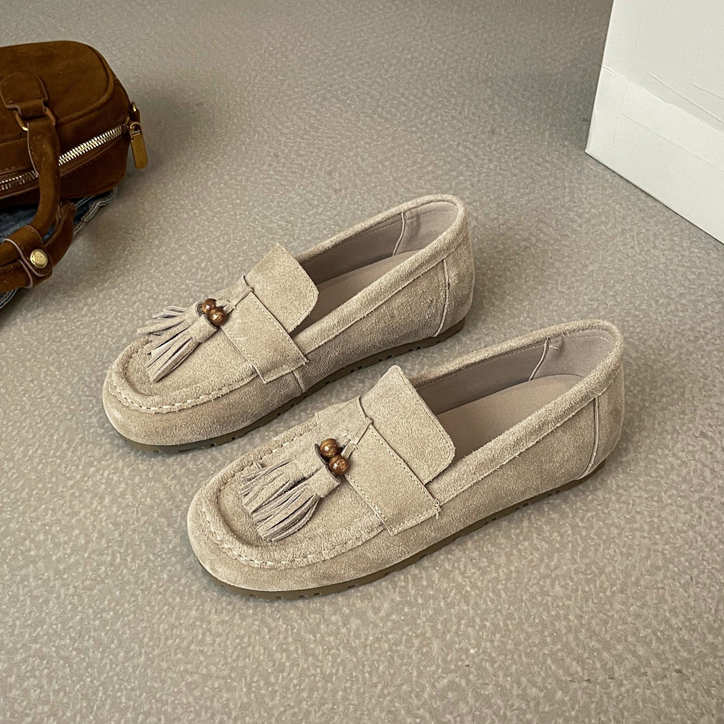 Beige women’s suede tassel flat loafers on carpeted floor next to brown suede handbag
