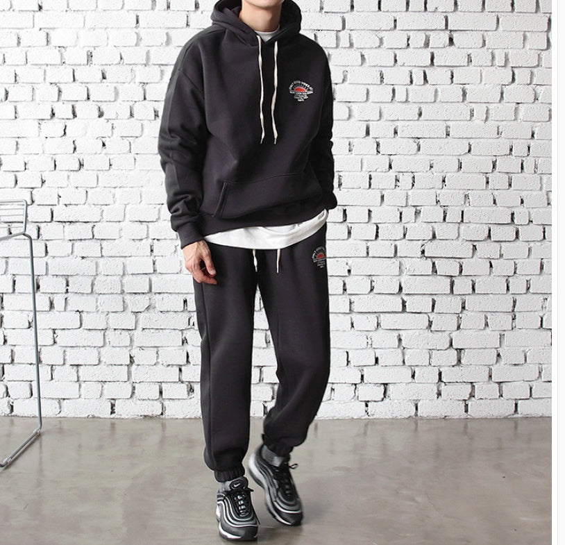 man wearing black hooded sportswear set with white drawstrings and sneakers against white brick wall