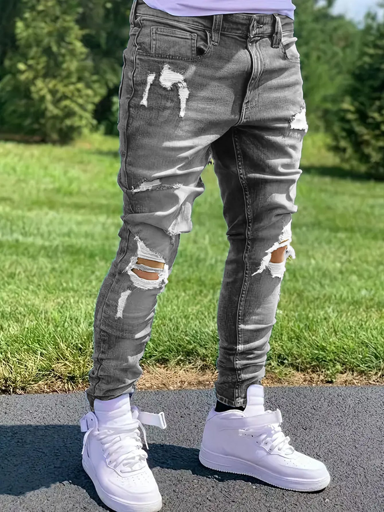 Man wearing ripped gray skinny jeans and white high-top sneakers standing outdoors on pavement with grass background