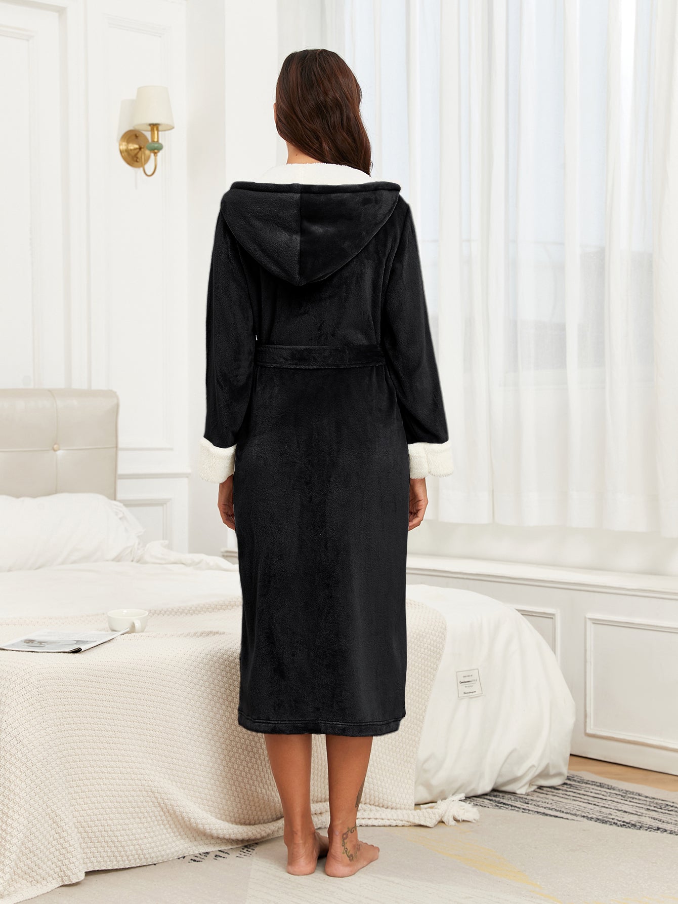 Woman wearing black plush fleece hooded robe with white cuffs standing barefoot in a bright bedroom