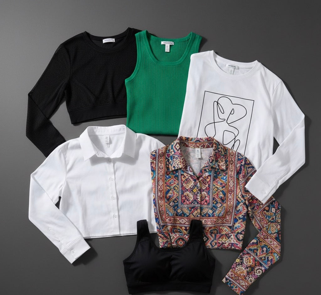 Flat lay of assorted women's tops including black crop sweater, green tank, white graphic tee, white button-up, patterned blouse, and black sports bra
