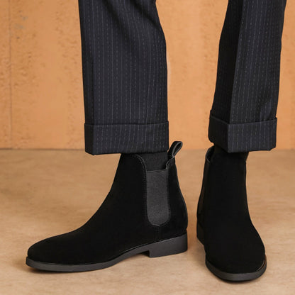 Black suede Chelsea boots with elastic side panels worn with pinstriped cuffed pants on brown floor