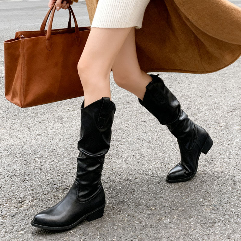 Woman walking on street wearing black leather mid-calf boots, beige coat, and carrying brown leather tote bag