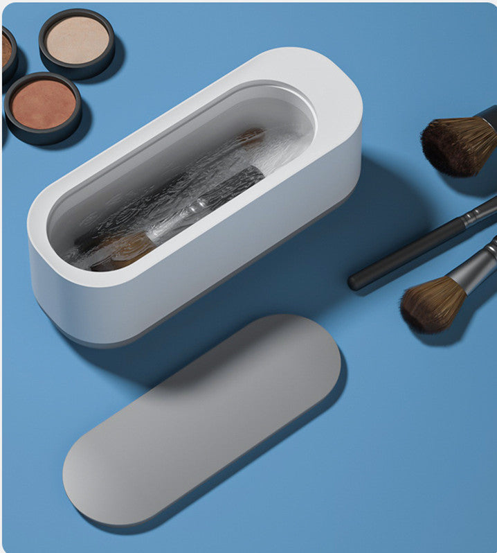 White ultrasonic makeup brush cleaner with brushes inside, surrounded by makeup brushes and powder compacts on blue surface