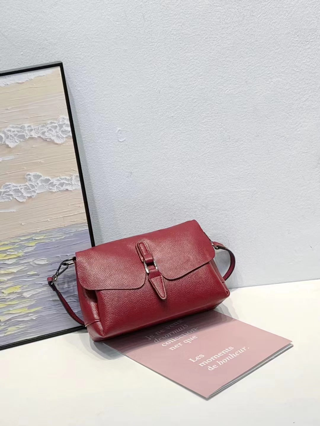 Red leather handbag with buckle detail on white table against gray wall, next to framed sea painting