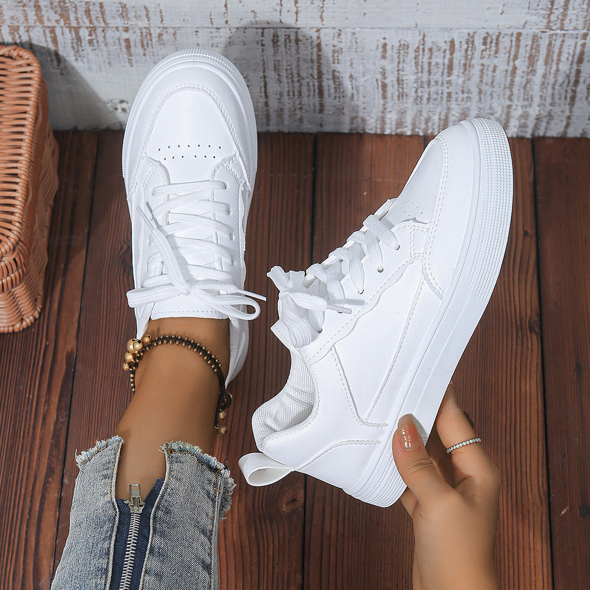Close-up of trendy white sneakers worn with blue frayed jeans and a beaded ankle bracelet on wooden floor
