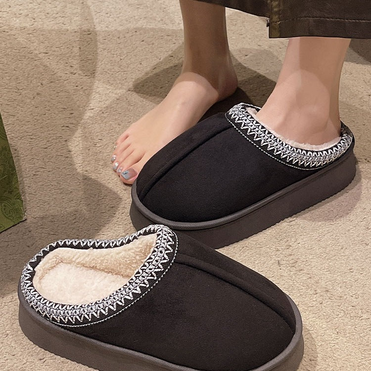 Black cozy slippers with white fur lining and decorative stitching worn by person on carpeted floor