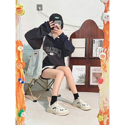 Person in black Chicago sweatshirt, black cap, white sneakers sitting on folding chair taking mirror selfie