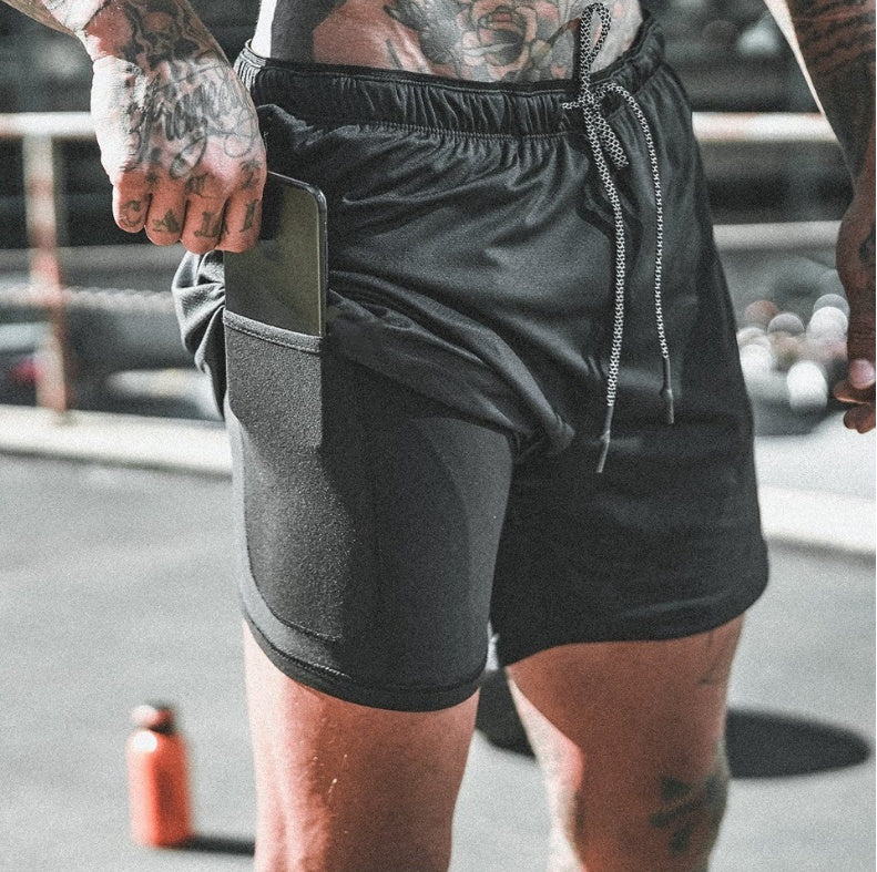 Close-up of tattooed man wearing black athletic shorts with side pocket holding a smartphone outdoors