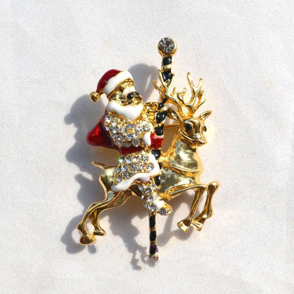 Gold-tone Santa Claus with red hat and rhinestones riding a gold reindeer brooch on white background