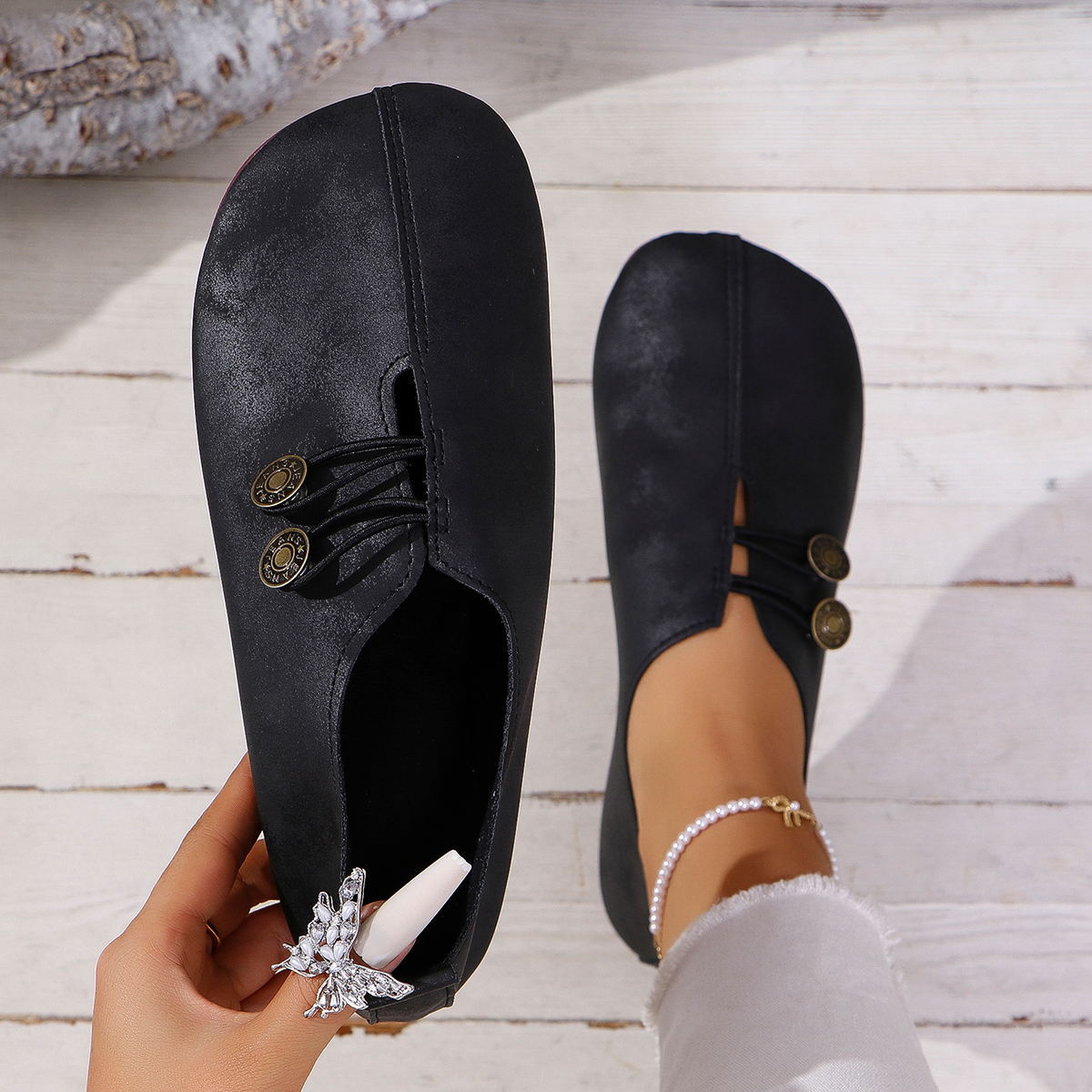 Black slip-on shoes with decorative buttons, worn with white pants and a pearl anklet on light wooden floor