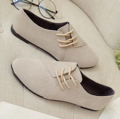 Pair of beige suede oxford shoes with light tan laces on a cushioned beige surface with round glasses nearby