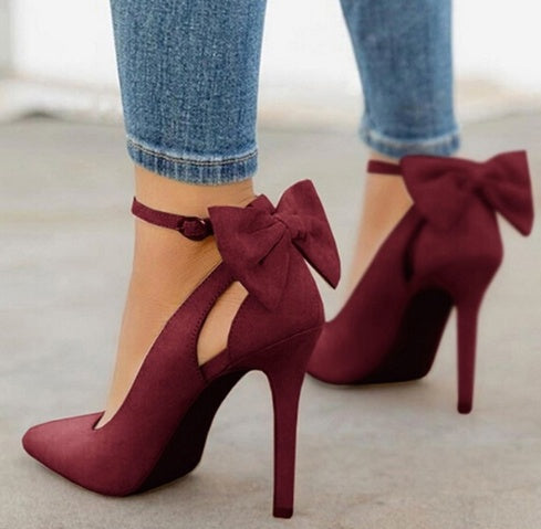 Close-up of woman wearing burgundy high heel shoes with large bow accents and ankle straps, paired with blue skinny jeans