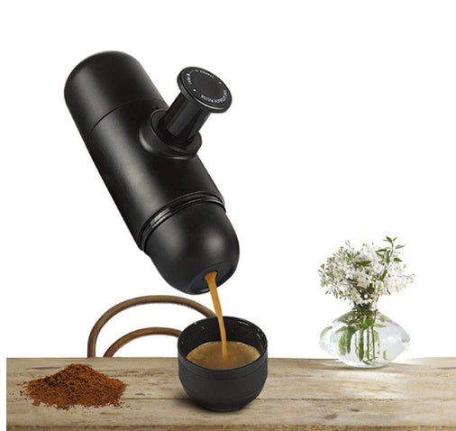Black portable espresso maker pouring fresh coffee into black cup on wooden table with coffee grounds and vase with white flowers