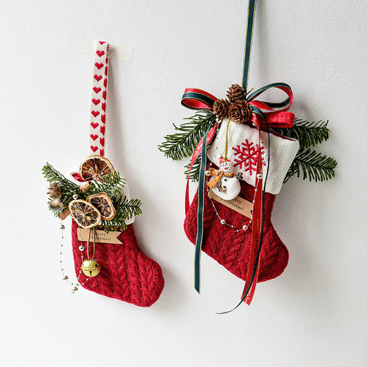 Two red knitted Christmas stockings decorated with pine branches, dried orange slices, pine cones, ribbons, and holiday charms hanging on a white wall