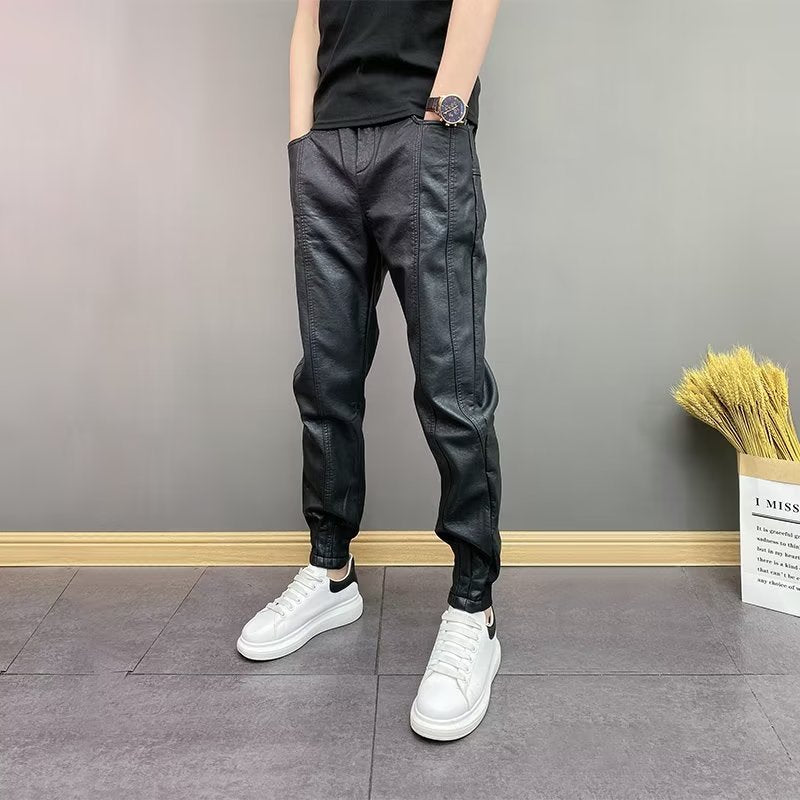 Man wearing black leather jogger pants and white sneakers standing against gray wall
