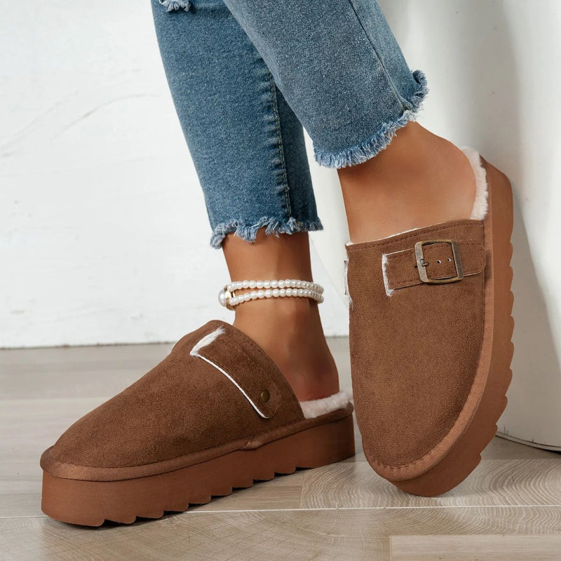 Brown suede slip-on platform slippers with white fur lining and buckle detail, worn with frayed hem jeans