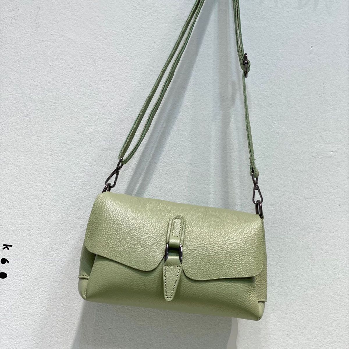 Light green textured leather crossbody bag hanging on wall with adjustable strap
