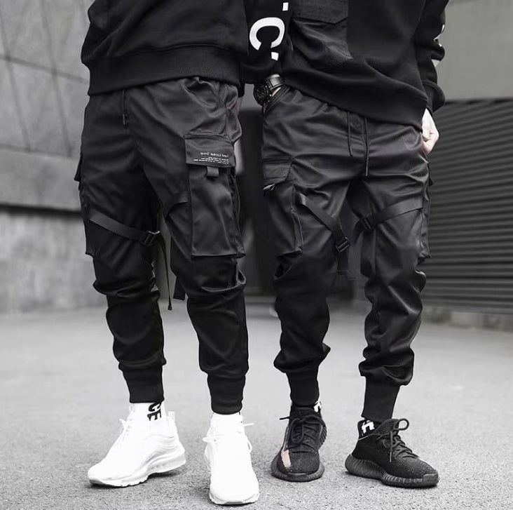 Two men wearing black cargo pants with multiple pockets and straps, paired with black and white sneakers in urban setting