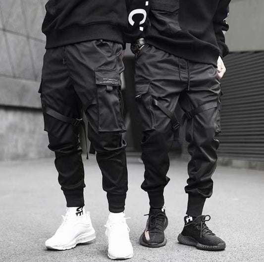 Two men wearing black cargo pants with multiple pockets and straps, paired with black and white sneakers in urban setting