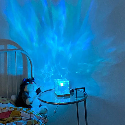 Blue LED cube night light on glass table casting ocean wave patterns, stuffed cat toy on bed nearby