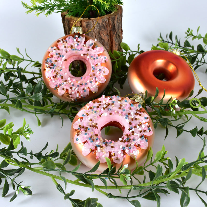 Three donut-shaped Christmas ornaments with pink icing and colorful sprinkles among green foliage