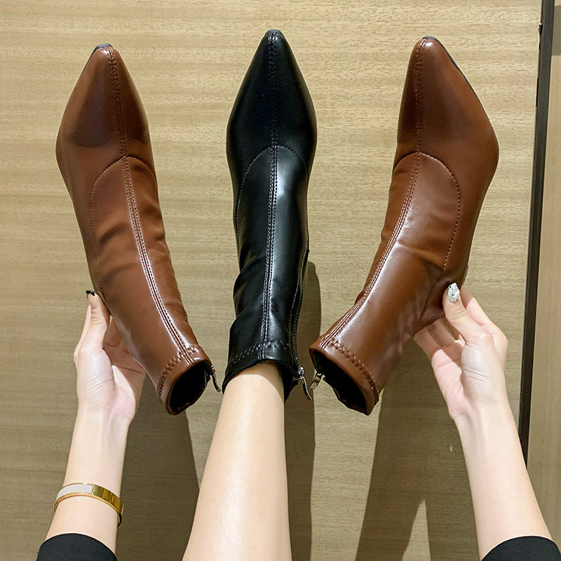 Pointed toe ankle boots in black worn on one foot and brown boots held by hands on wood background