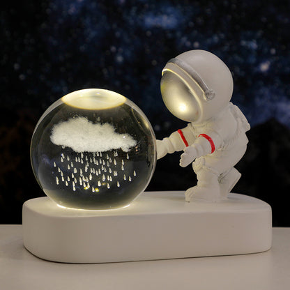 White astronaut figurine reaching towards illuminated glass globe with etched rain cloud on white base