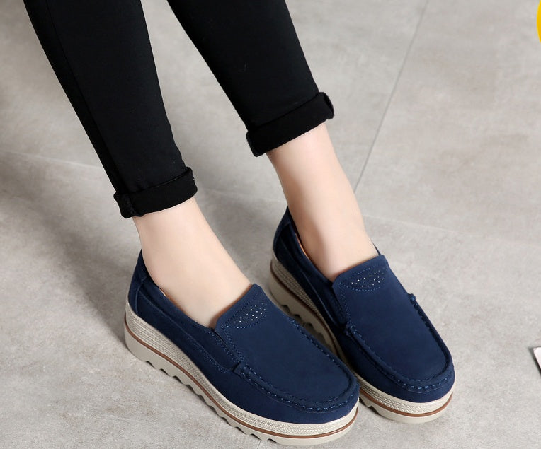 Blue slip-on platform shoes worn with black rolled-up pants on a gray floor