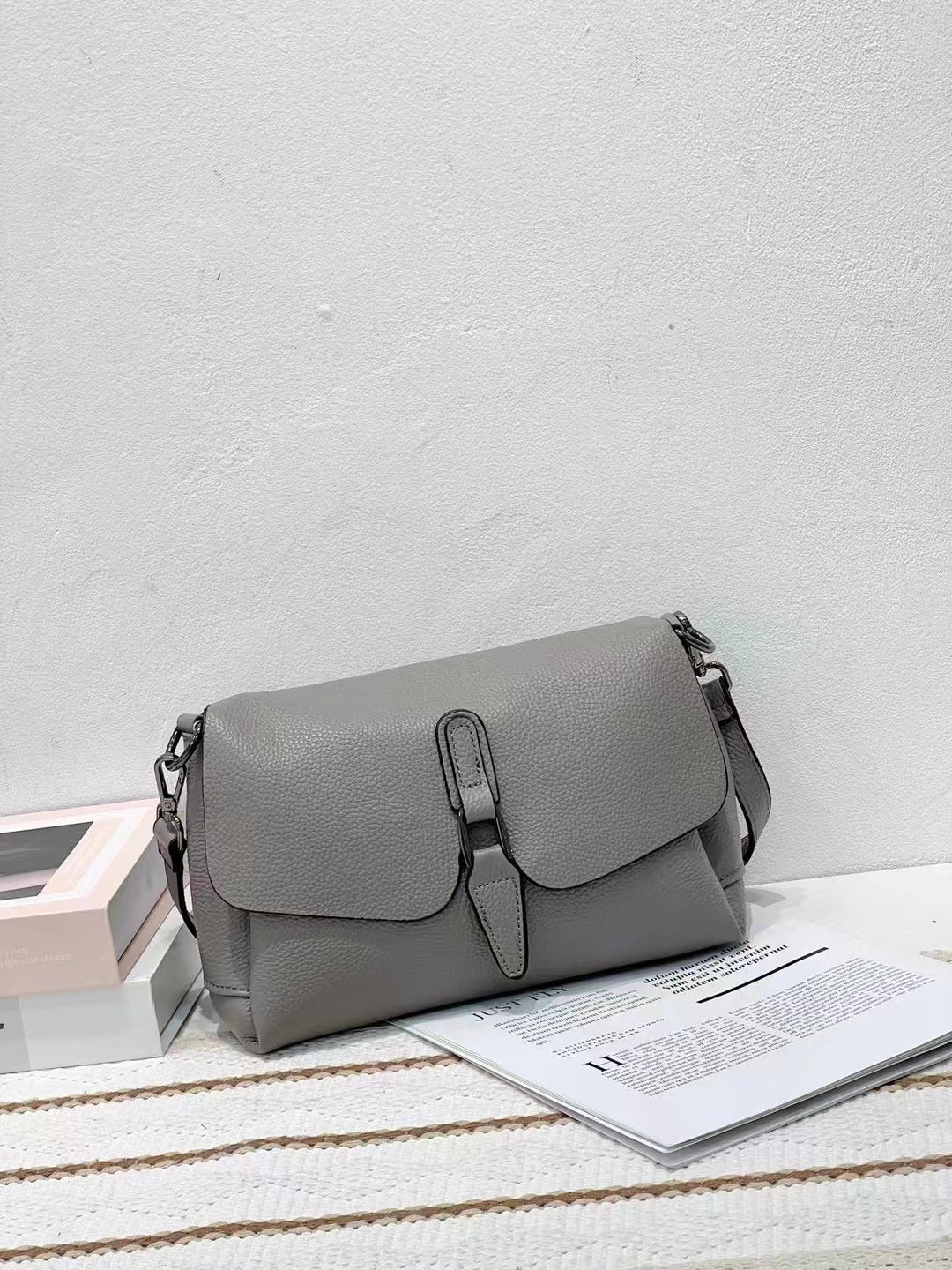 Gray leather crossbody handbag on striped fabric with open magazine and stacked books