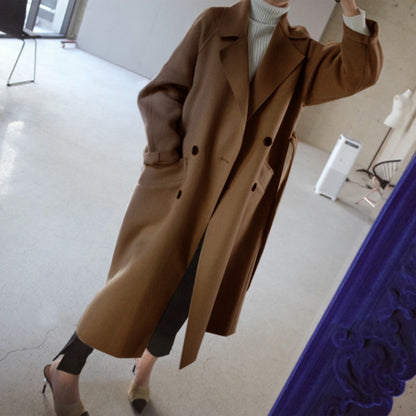 Woman wearing long camel coat, white turtleneck, black pants, and beige heels in minimal room