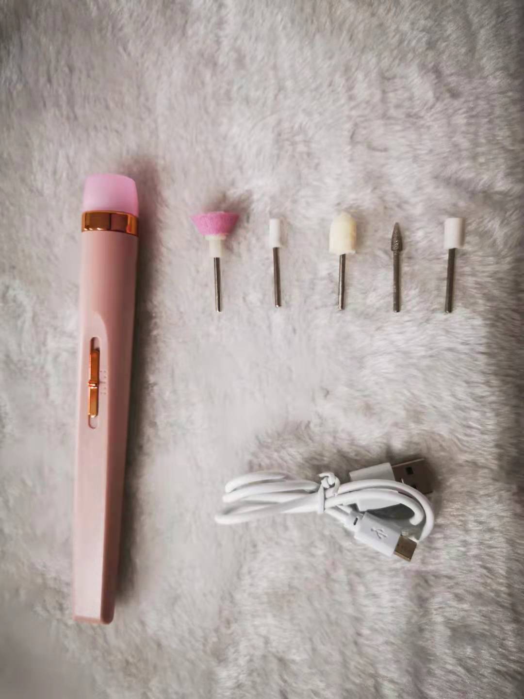 Pink electric nail drill kit with six metal and foam drill bits and a white USB charging cable on gray fabric