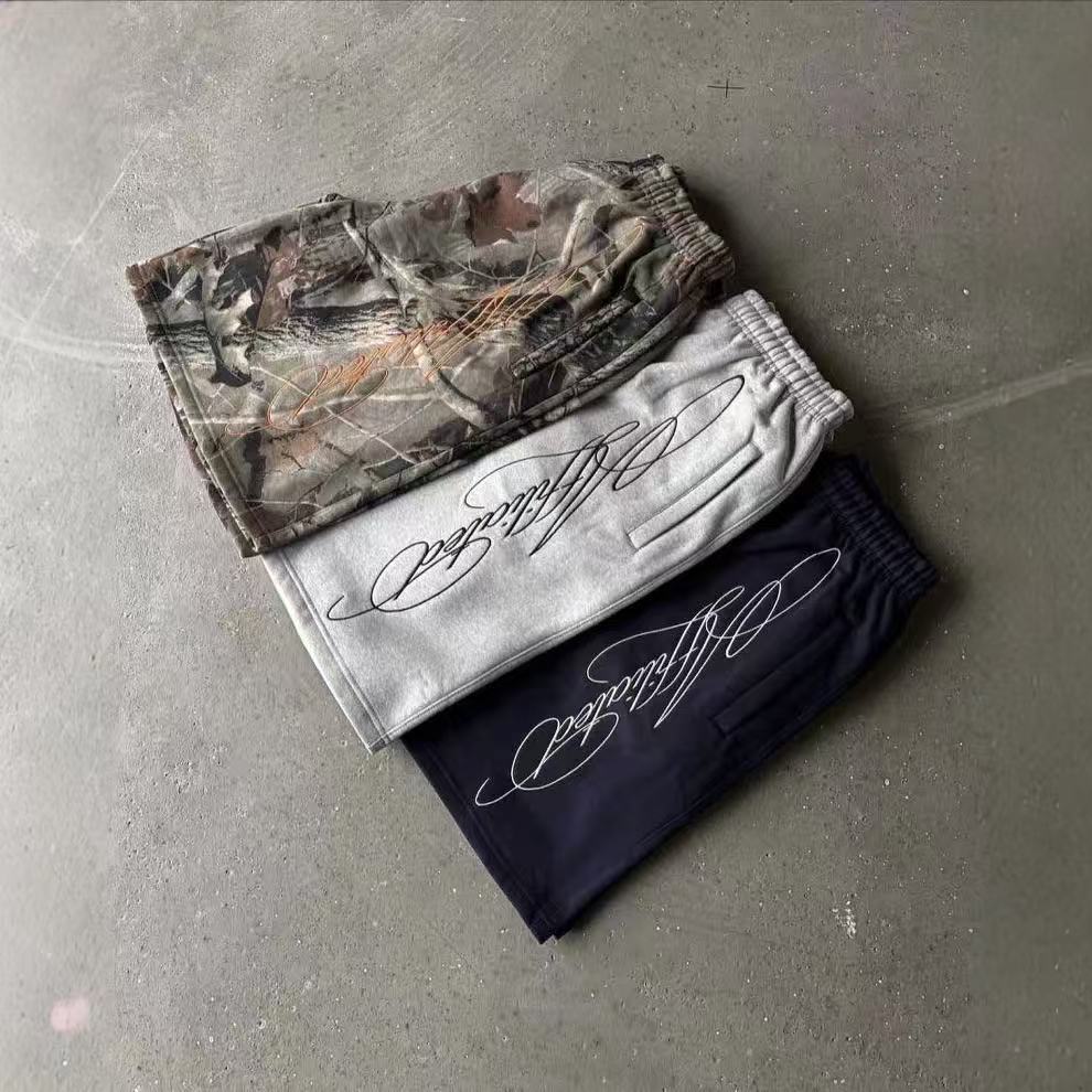 Three folded sweatpants in camouflage, gray, and black with elegant script text on a concrete floor