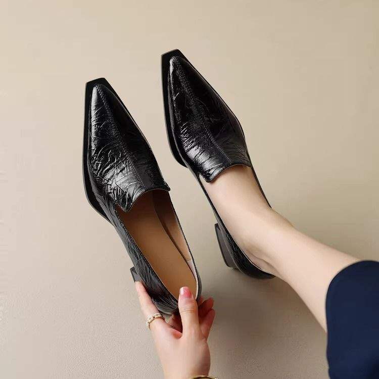 Black pointed toe leather loafers worn and held in hand on beige background