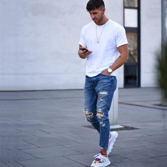 Man in white t-shirt and ripped jeans walking on pavement while using smartphone outdoors