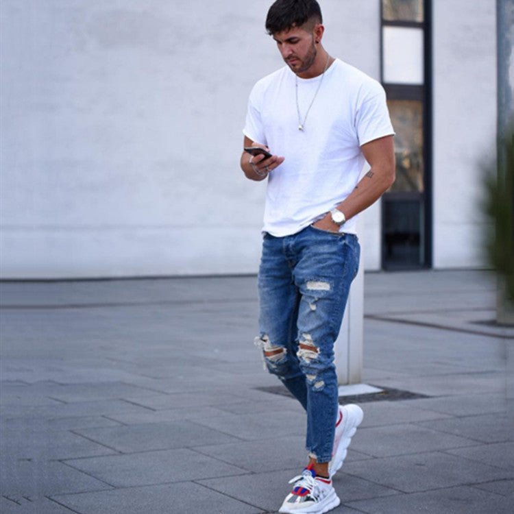 Man wearing white t-shirt and ripped blue jeans walking outdoors while looking at phone