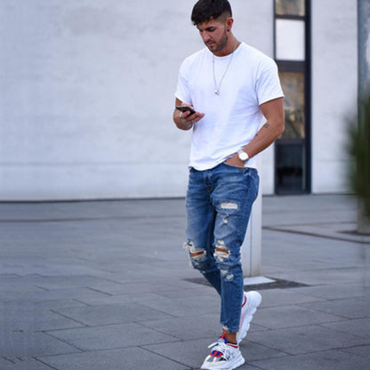 Man wearing white t-shirt and ripped blue jeans walking outdoors while looking at phone