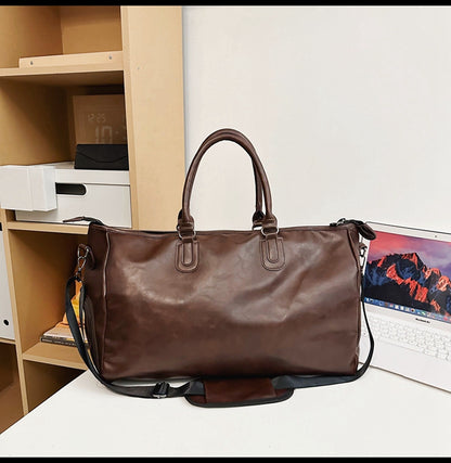 Brown leather travel duffel bag with handles and adjustable shoulder strap on white desk near bookshelf and laptop