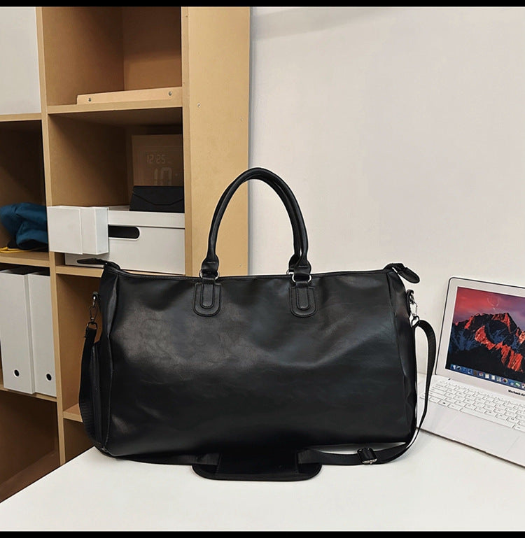 Black leather duffel bag with handles and shoulder strap on white desk next to open laptop and wooden shelves
