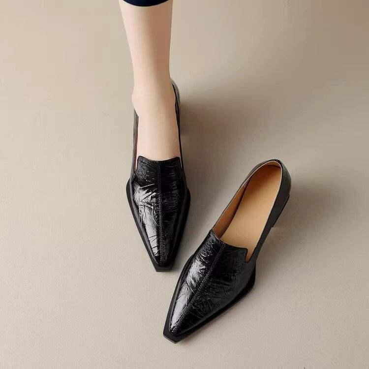 Black pointed-toe leather loafers on light floor, one worn on foot, sleek modern style
