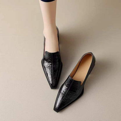 Black pointed-toe leather loafers on light floor, one worn on foot, sleek modern style
