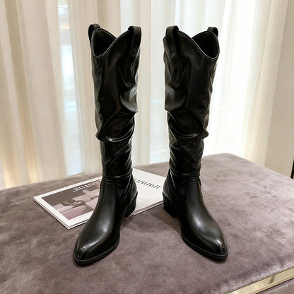 Pair of black knee-high leather boots with pointed toes on a gray soft surface near sheer curtains