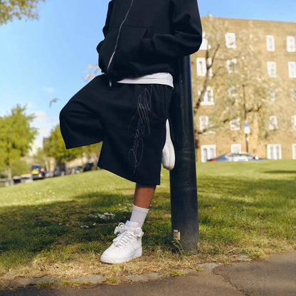 Person in black hoodie and black shorts with script logo leaning on pole in park wearing white sneakers