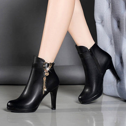 Black leather high heel ankle boots with gold zipper charm on model's feet against grey background