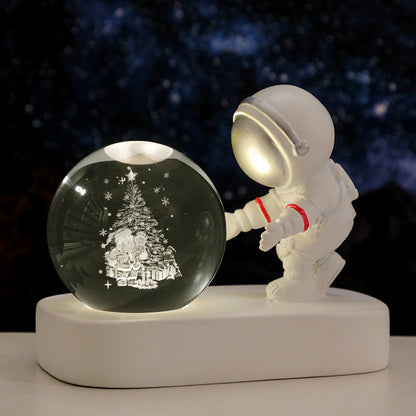 White astronaut figurine reaching towards illuminated Christmas snow globe with Santa and tree