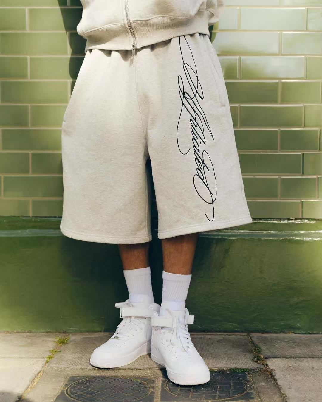 Person wearing beige shorts with black script print, white socks, and white high-top sneakers against green tiled wall